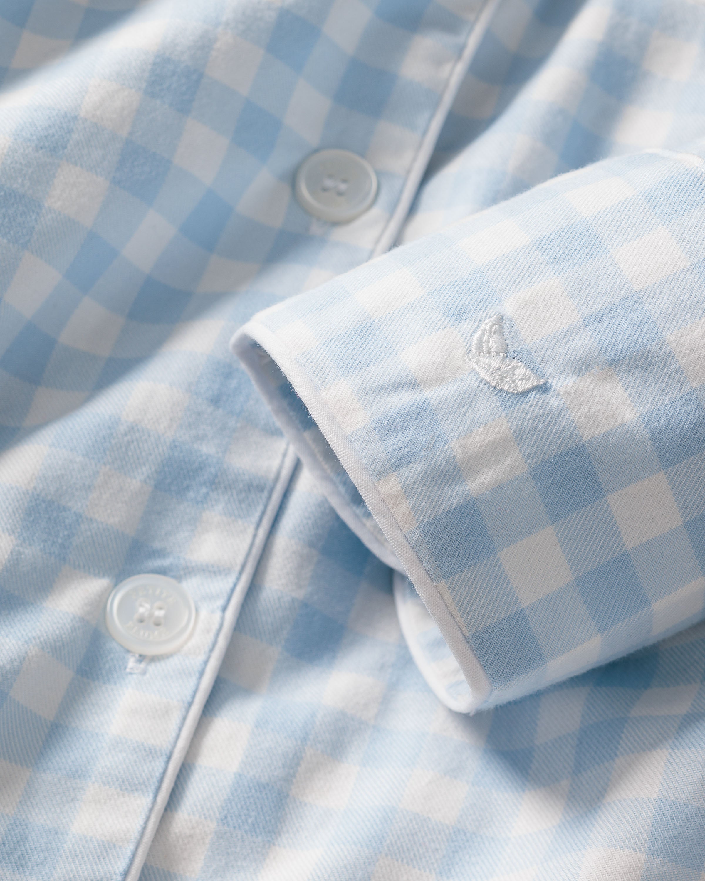 Close-up of Petite Plumes Womens Twill Pajama Long Sleeve Short Set in light blue gingham, featuring two white buttons and part of the cuff. The soft cotton twill fabric offers a cozy feel, with subtle embroidery enhancing its charm.