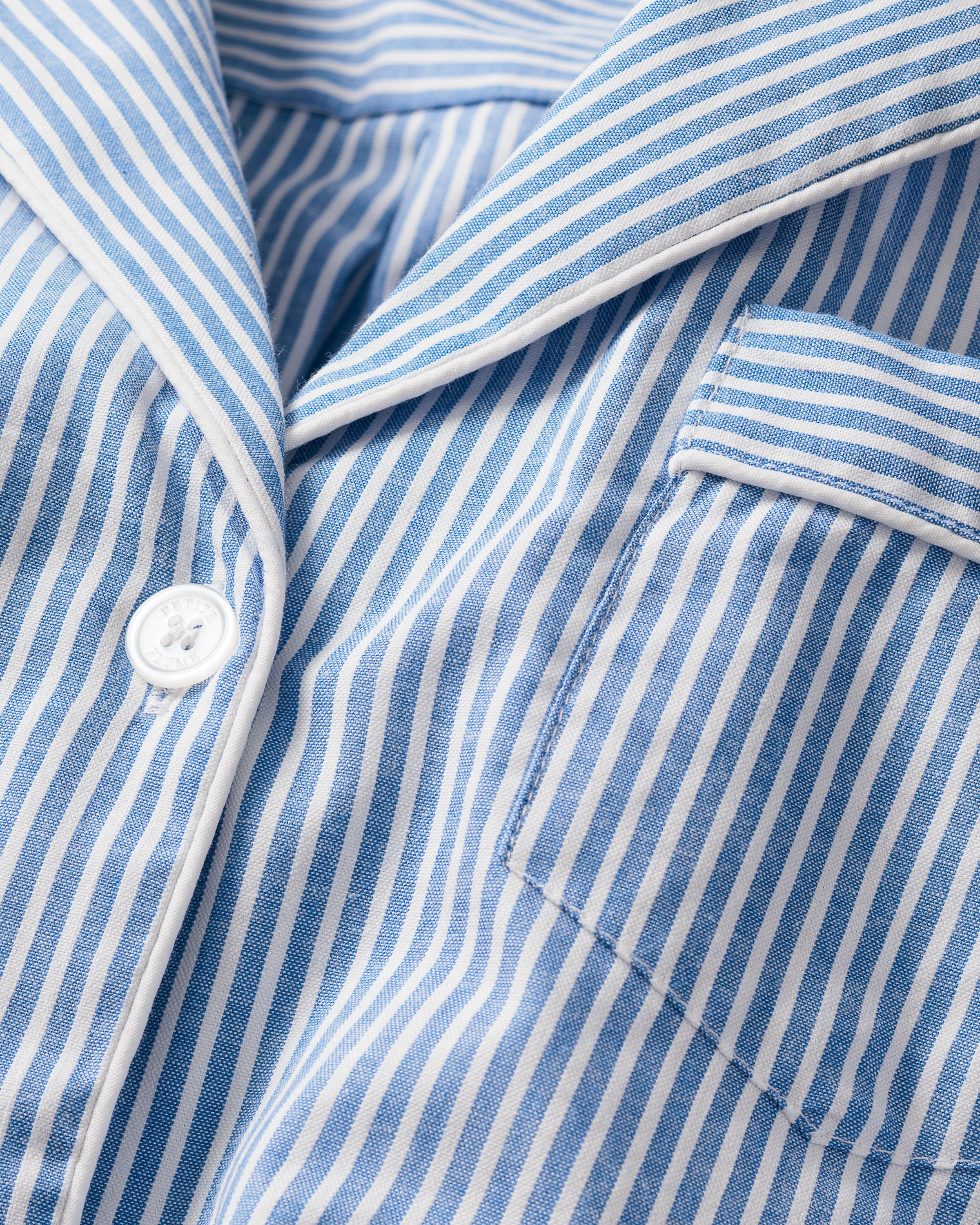 Close-up of Womens Twill Long Sleeve Short Set in French Blue Seersucker from Petite Plume—featuring a blue and white striped shirt with collar, button, right-side pocket, made from lightweight 100% cotton fabric reminiscent of classic seersucker pajamas.