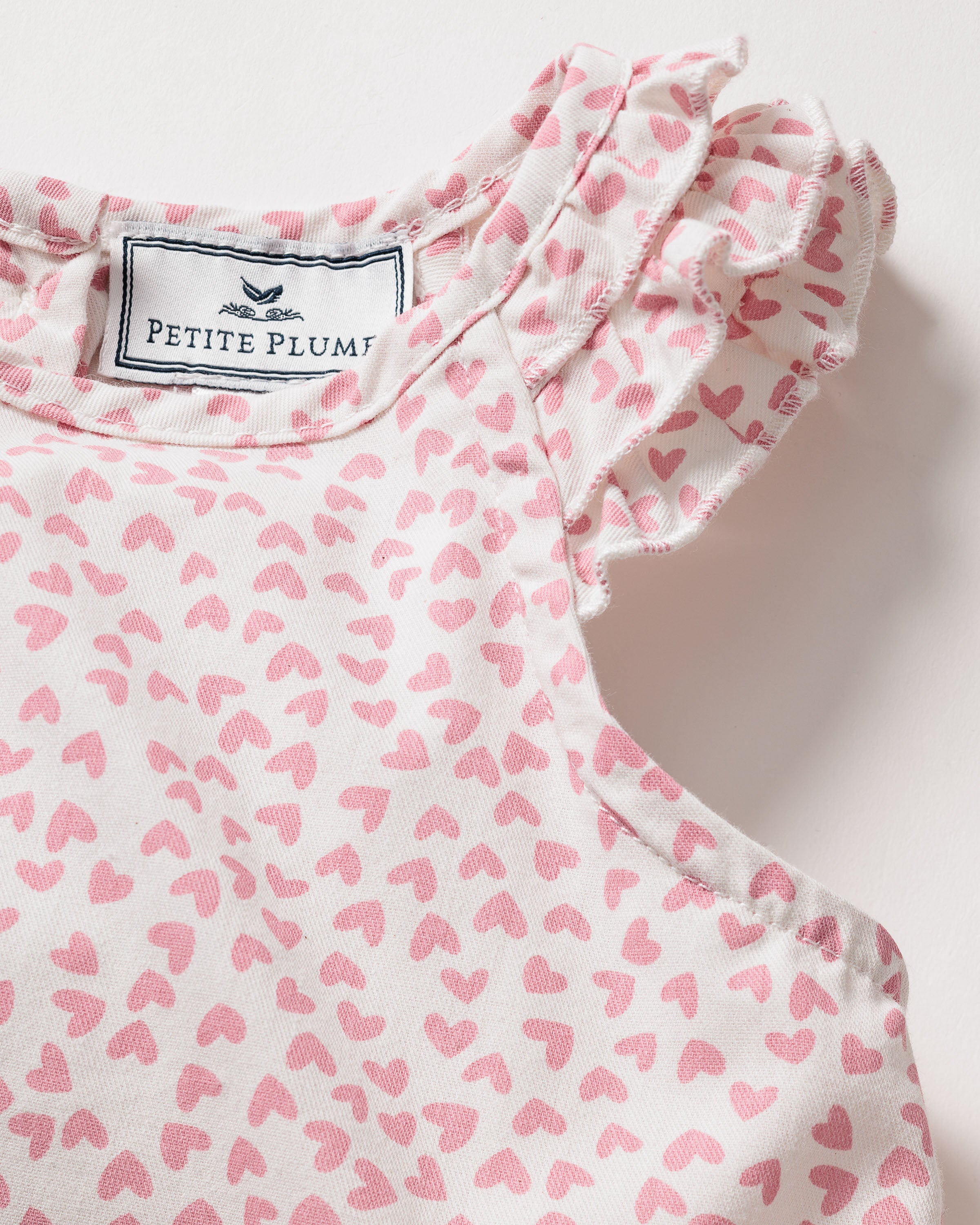 A close-up of the Girls Twill Amelie Nightgown in Sweethearts shows a sleeveless white dress with pink hearts, made from lightweight woven cotton. Ruffled shoulders add luxury sleepwear vibes. The label Petite Plume is visible inside the neckline.