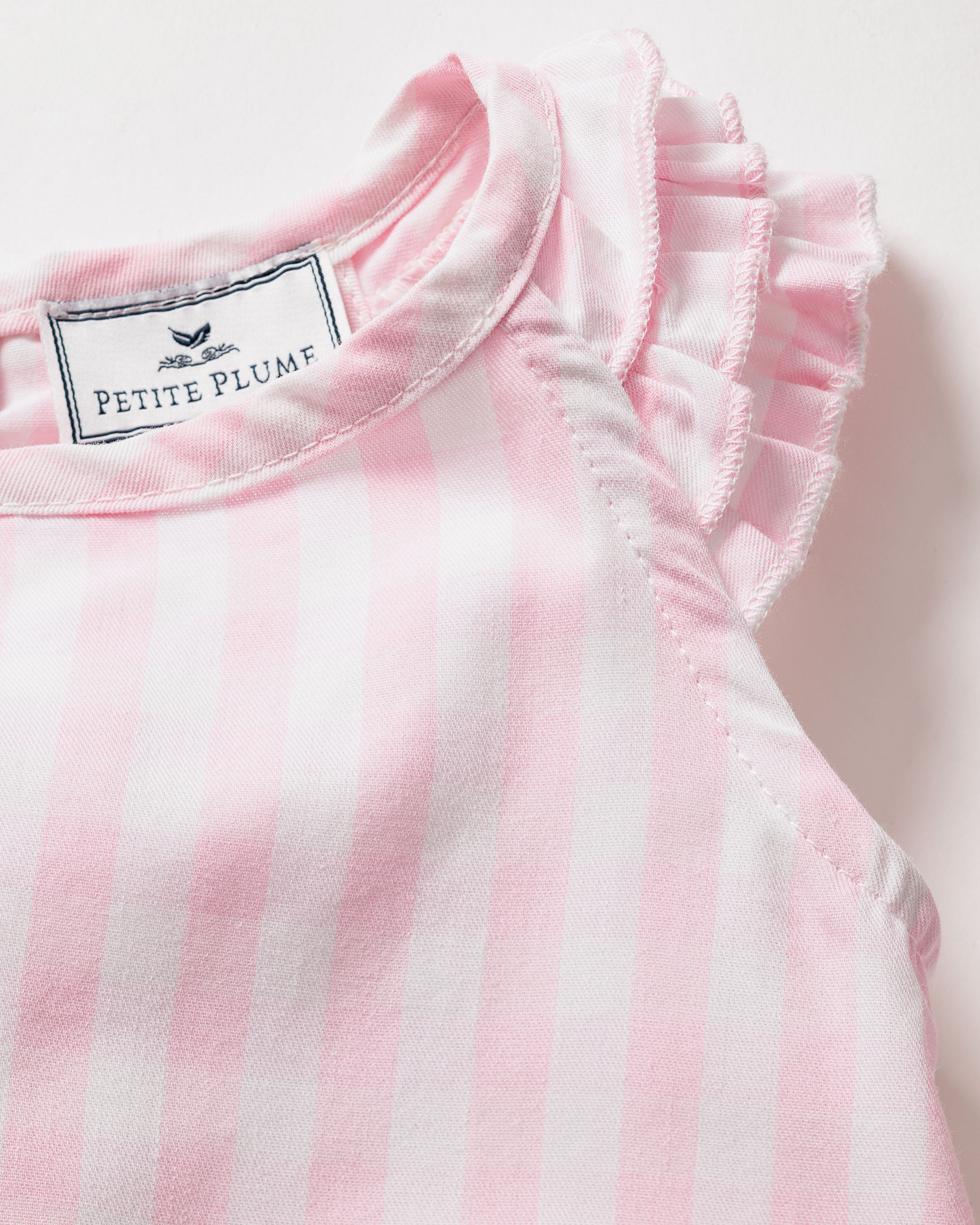A close-up of a pink gingham garment with a ruffled sleeve, made from yarn-dyed cotton marked Petite Plume with a bird logo. The fabric feels soft and lightweight, epitomizing luxurious sleepwear. This is the Girls Twill Amelie Short Set by Petite Plume.
