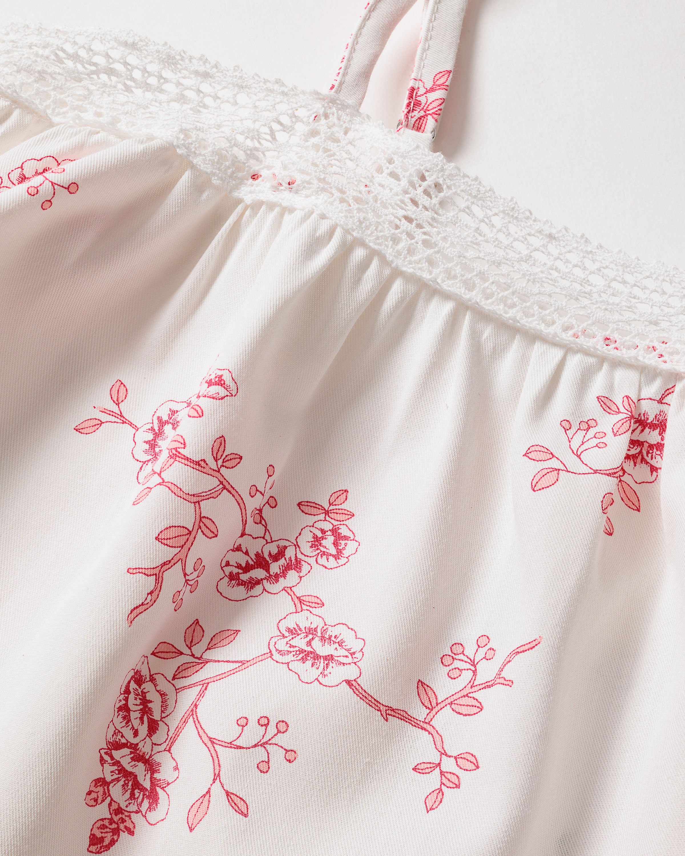 A close-up of the Petite Plume Girls Twill Lily Nightgown in English Rose Floral, showcasing its white fabric adorned with red floral designs. Featuring intricate lace along the top edge and two thin straps, this cotton sleepwear exudes elegance with stylized flowers and leaves.