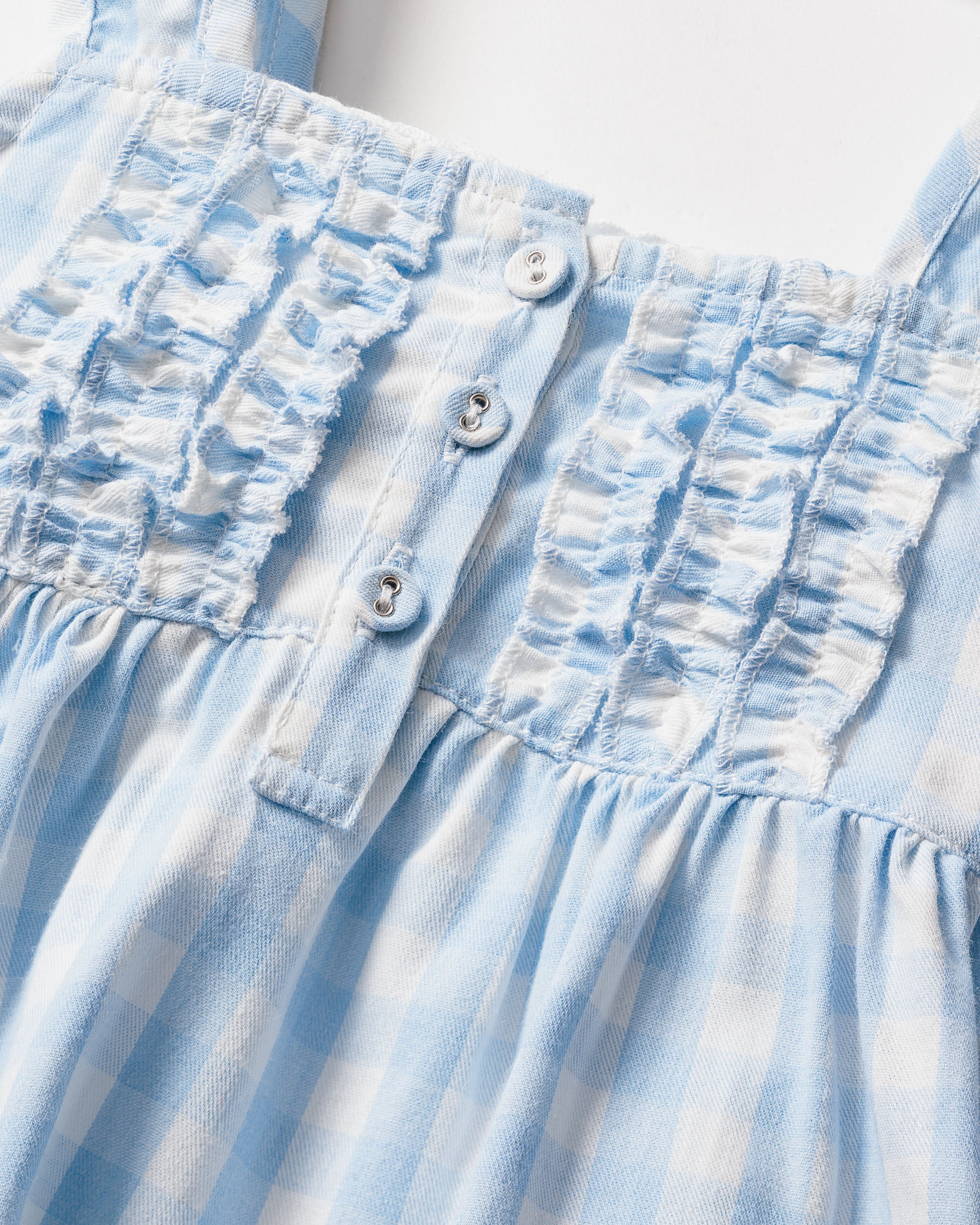 Close-up of Petite Plumes Girls Twill Charlotte Nightgown in Light Blue Gingham, highlighting its smocked bodice, three white buttons, soft textured fabric, vertical ruffles, and thin shoulder straps for a charming look.