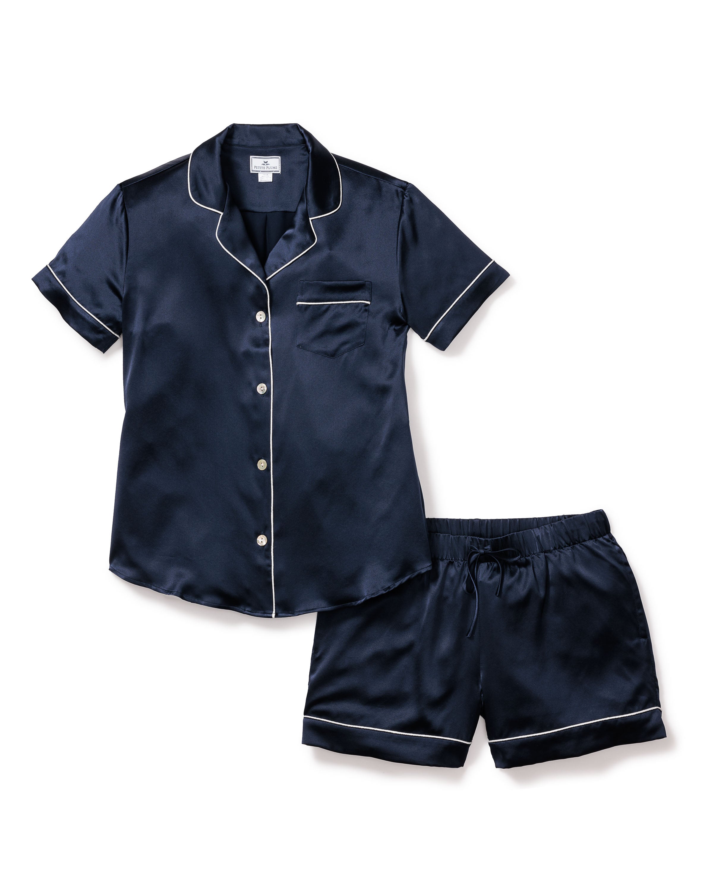 The Womens Silk Short Set in Navy by Petite Plume is a luxurious sleepwear ensemble, featuring a short-sleeved button-up top with white piping on the collar, sleeves, and pocket. The matching shorts have an elastic waistband and white piping on the hems for a restful night.