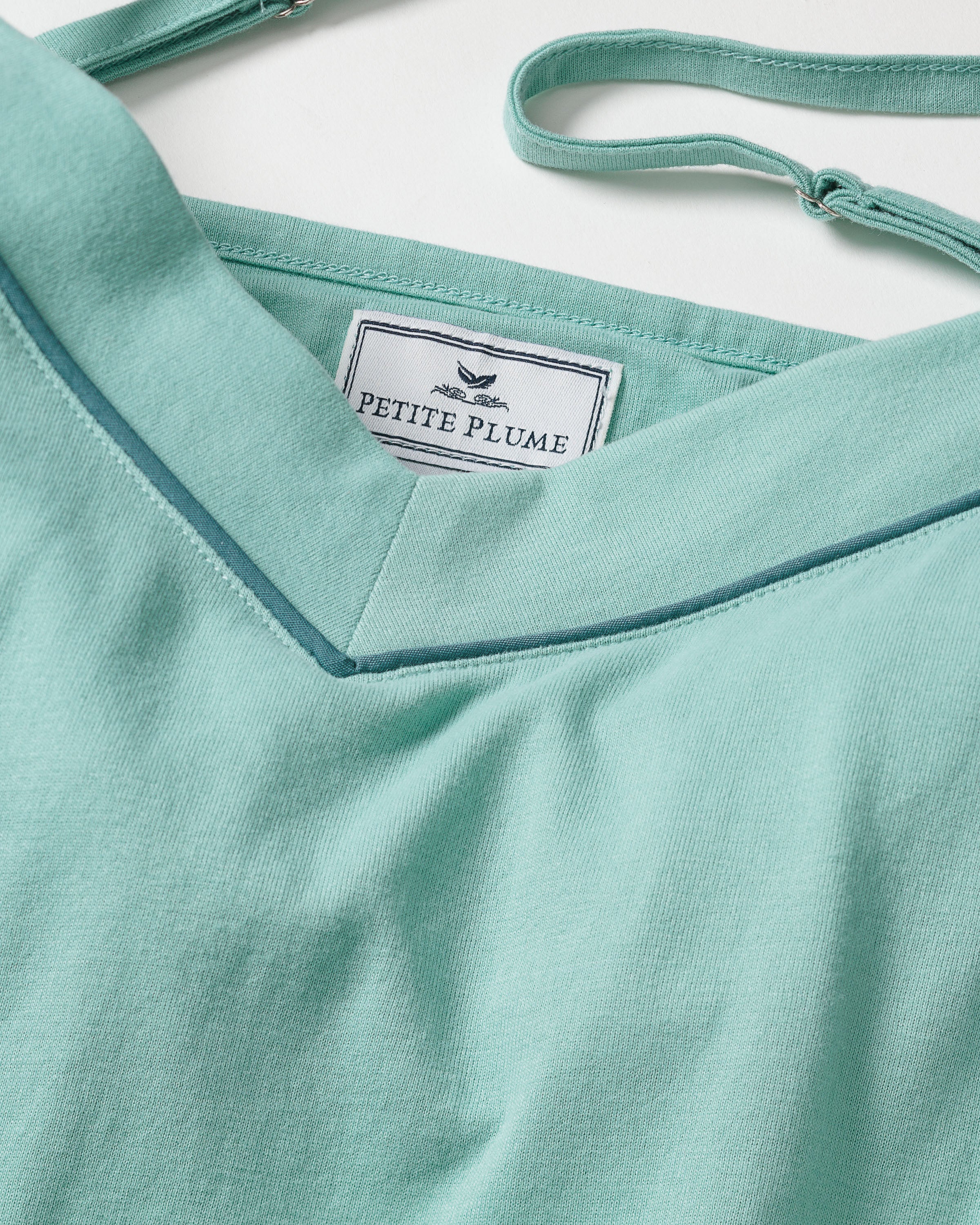 A close-up of the Womens Pima Sylvie Cami Short Set in Seaglass reveals a smooth, neatly stitched teal fabric crafted from soft Pima cotton. Inside the neckline, the Petite Plume tag is visible, hinting at its elegant pajama set design with an intriguing strap detail.