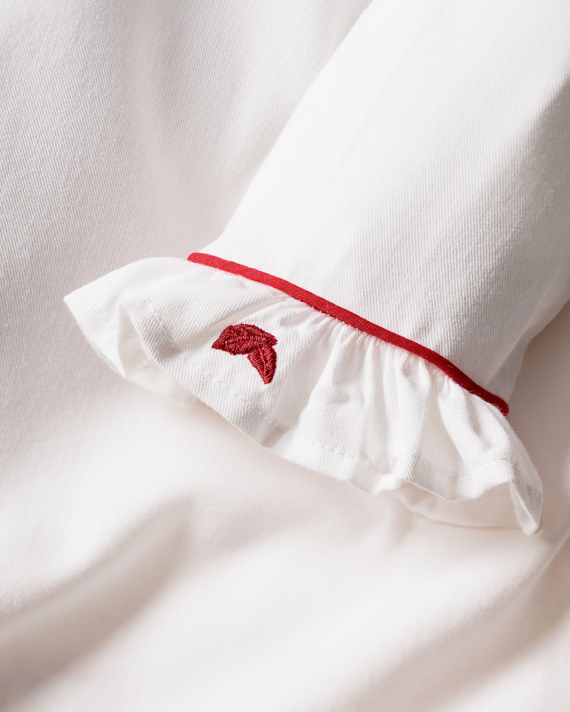 The Girls White Sophia Nightgown by Petite Plume features a close-up of a ruffled cuff with red piping, adorned with an embroidered heart. Made from soft, textured cotton fabric, it creates a cozy and elegant appearance.