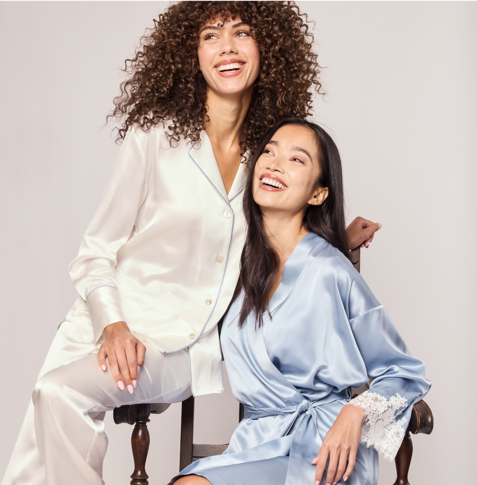 Two women in white silk and light blue pajamas seated together smiling