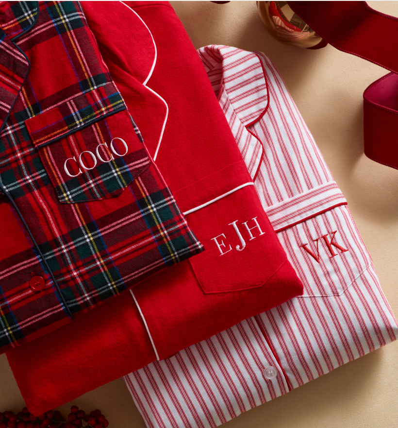 Three sets of folded pajamas with monogrammed initials on the pockets. One pair is plaid with COCO, one is solid red with EJH, and one is red-and-white striped with WK. Red ribbon is nearby. Three sets of folded pajamas with monogrammed initials on the pockets. One pair is plaid with COCO, one is solid red with EJH, and one is red-and-white striped with WK. Red ribbon is nearby.