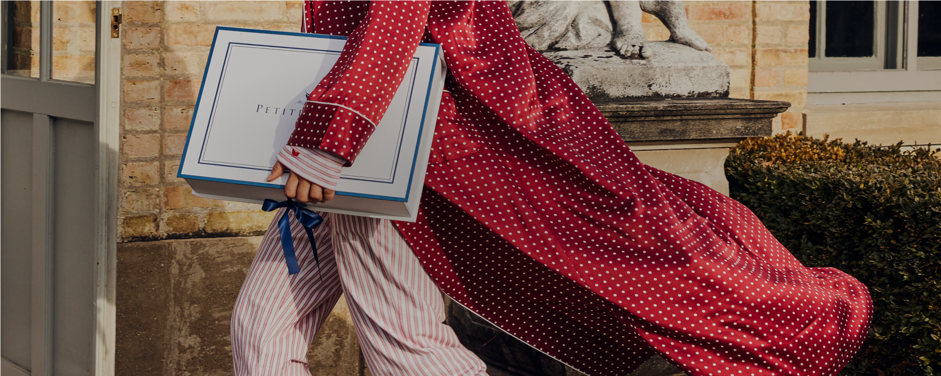 A person wearing pink and red polka dot pajamas walks outdoors holding a large white box with a blue border and ribbon. A stone statue and trimmed bushes are visible in the background. A person wearing pink and red polka dot pajamas walks outdoors holding a large white box with a blue border and ribbon. A stone statue and trimmed bushes are visible in the background.