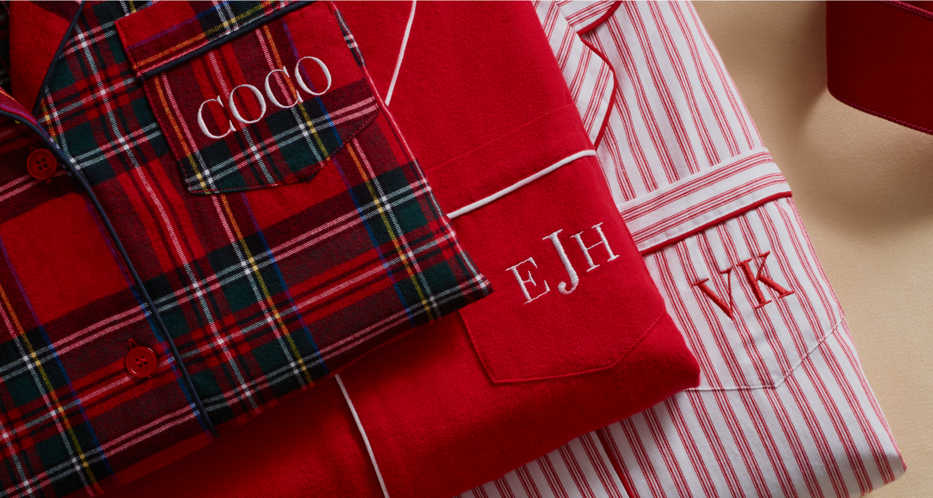 Three sets of folded pajamas with monogrammed names and initials: COCO on red plaid, EJH on solid red, and VK on pink and white stripes, arranged side by side on a beige surface. Three sets of folded pajamas with monogrammed names and initials: COCO on red plaid, EJH on solid red, and VK on pink and white stripes, arranged side by side on a beige surface.