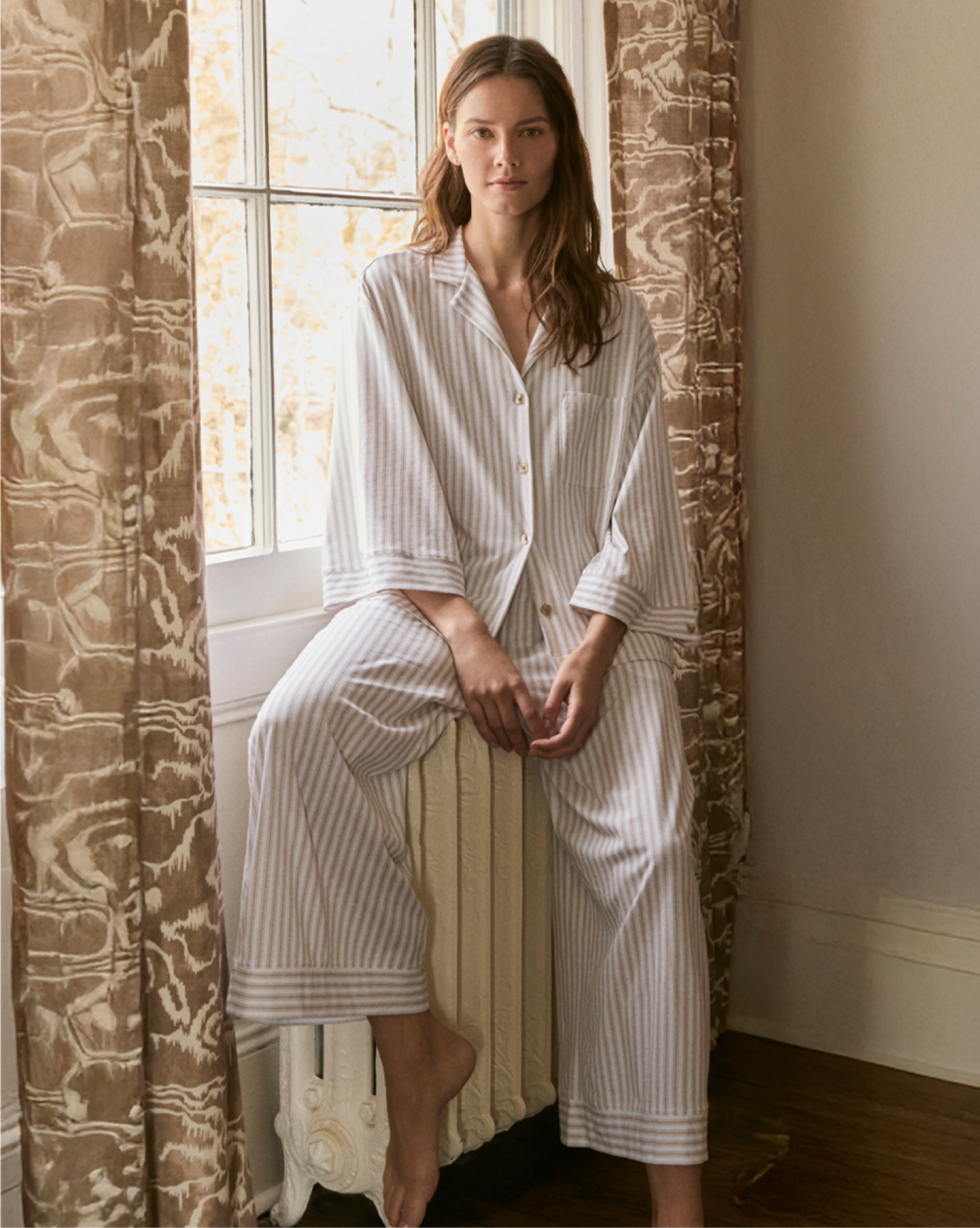 A woman in loose, light-striped pajamas sits on a radiator by a window, framed by brown patterned curtains, in a softly lit room with wooden floors.