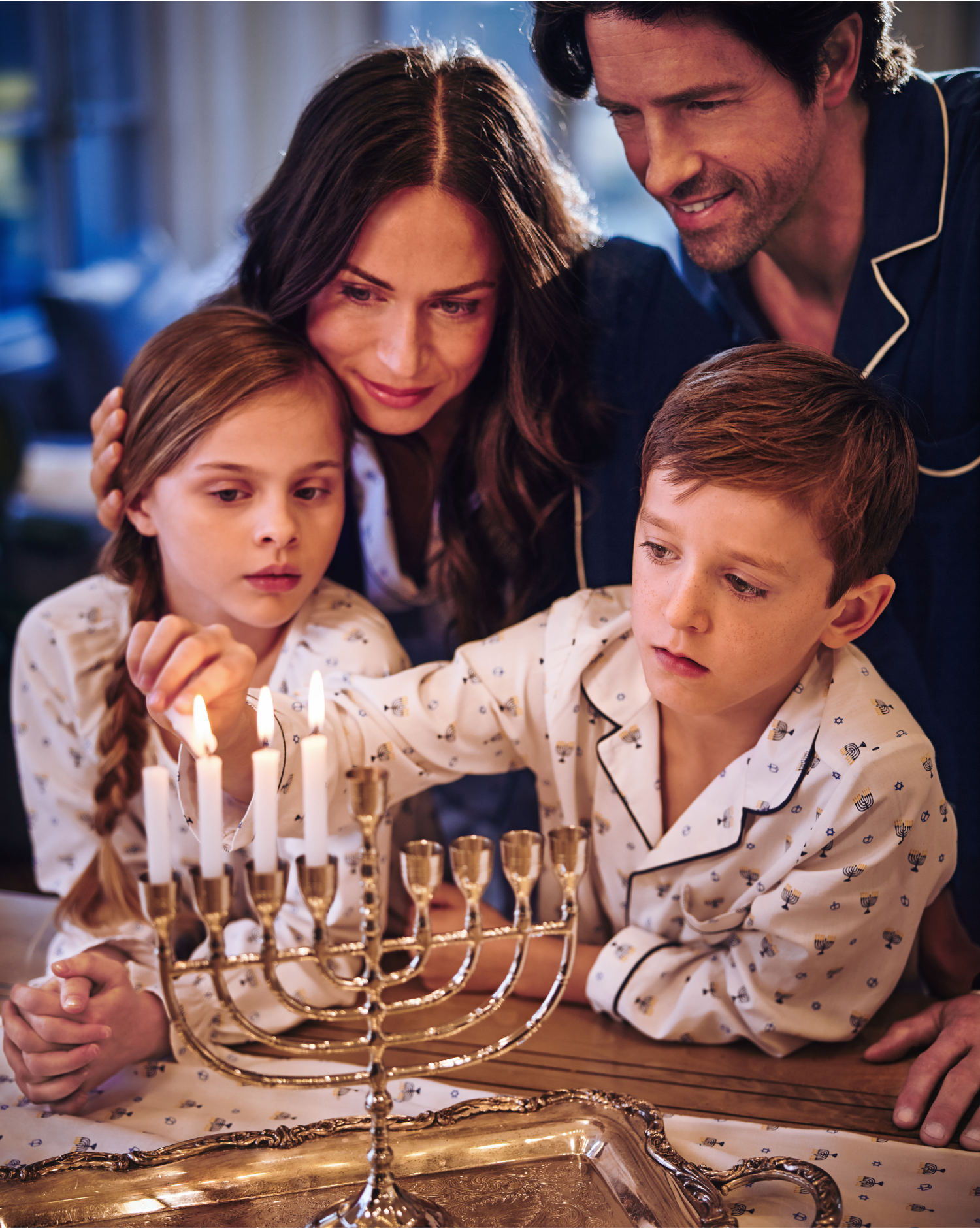A family of four, wearing pajamas, gathers around a menorah. The young boy lights the candles while the girl, mother, and father watch closely, smiling warmly, celebrating Hanukkah together at home.