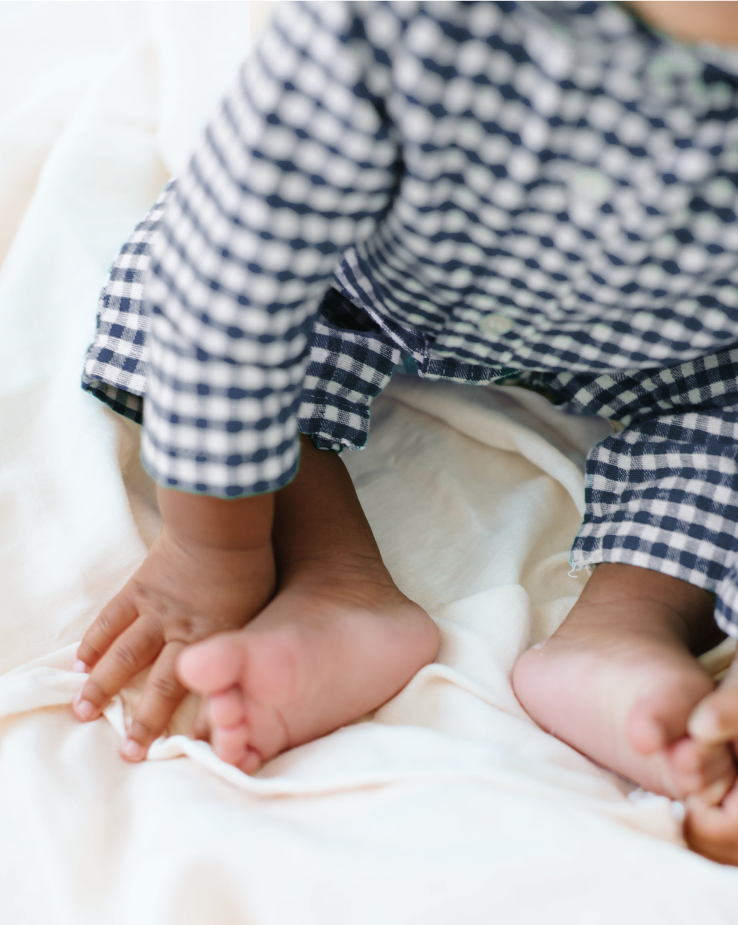A baby wearing a blue and white checkered outfit sits on a soft, cream-colored blanket with bare feet visible.