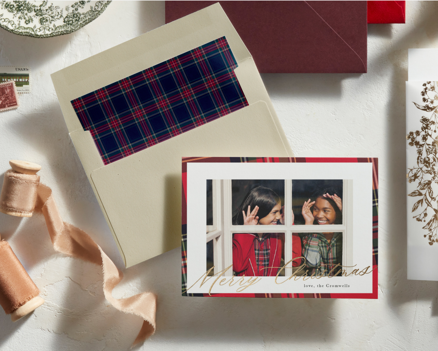 A holiday card on a table shows two smiling children looking out a window. The card reads Merry Christmas in gold script. Nearby are an envelope with a plaid liner, ribbon spools, and other cards.