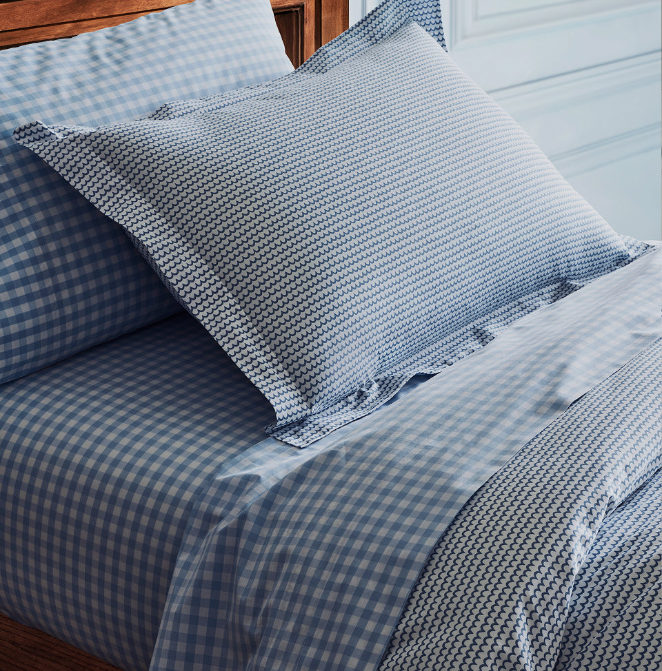 A neatly made bed with blue and white gingham-patterned sheets and pillowcase, and a decorative pillow featuring a small blue geometric pattern, against a wooden headboard and light-colored wall.