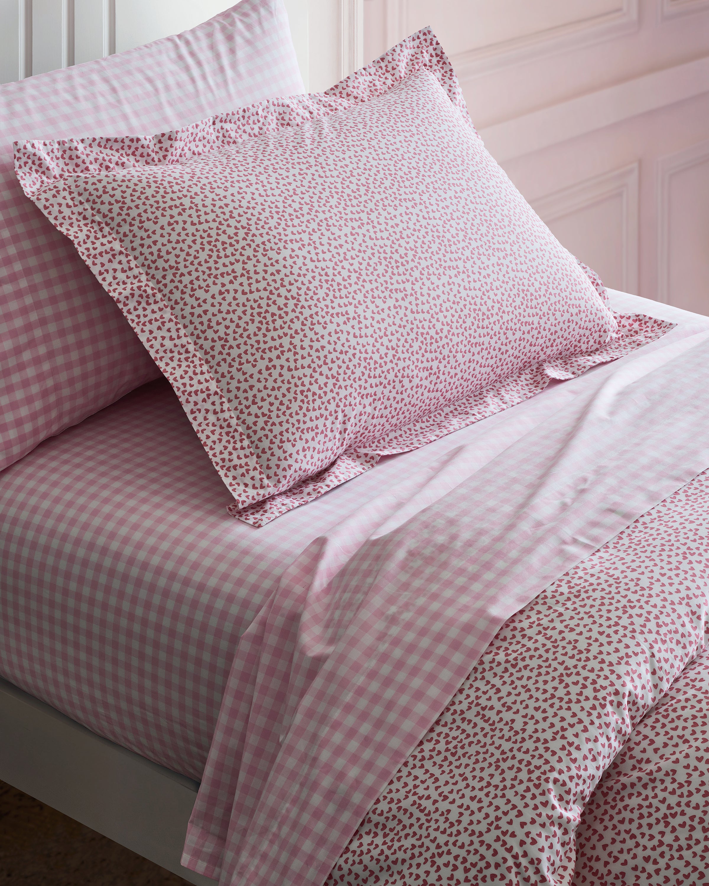 The Luxe Premium Cotton Pink Gingham Bed Sheets from Petite Plume feature a mix of floral and checkered patterns on luxurious long-staple cotton. A floral, frilled-edge pillow complements the checkered percale sheet, with light-colored wall paneling in the background.