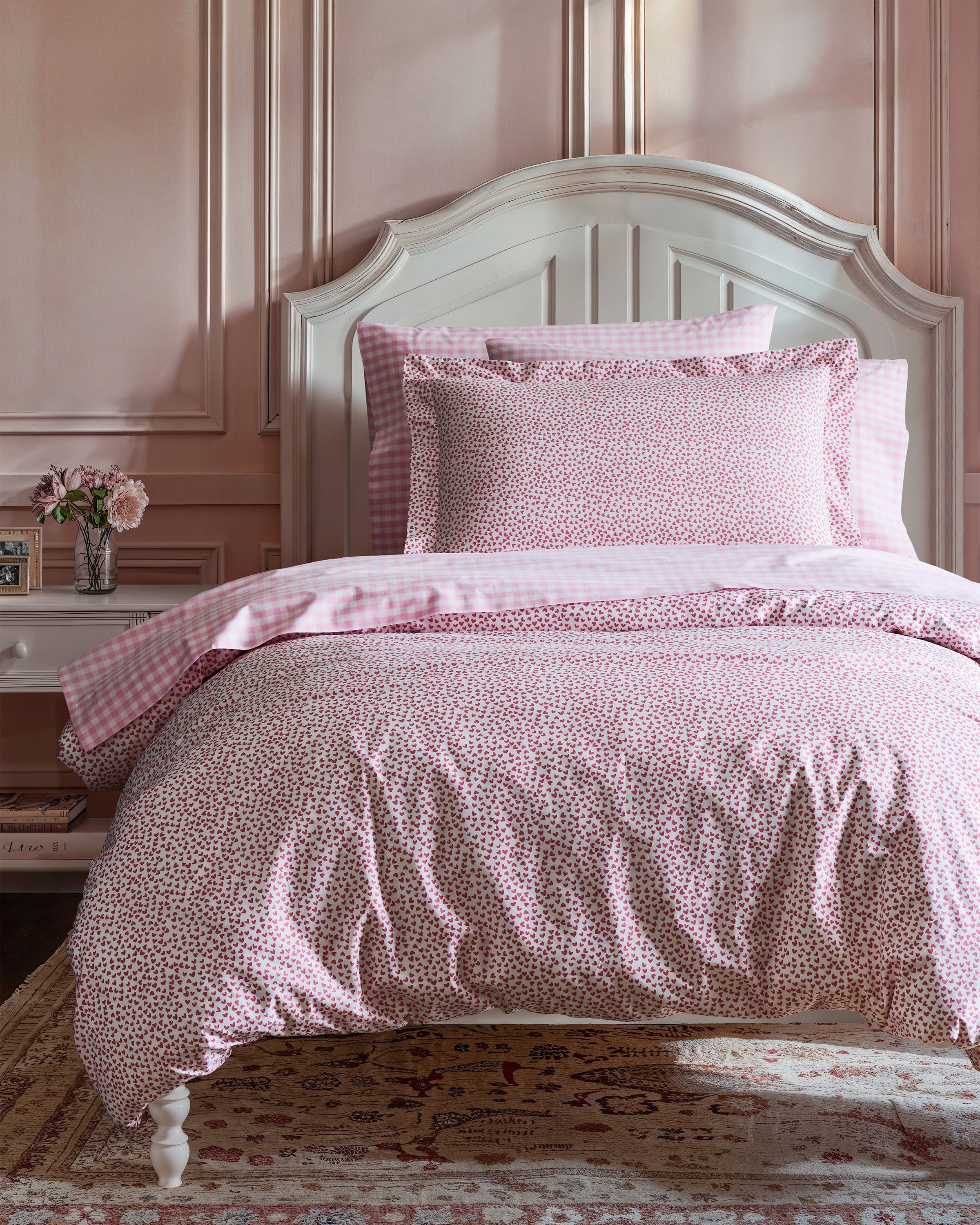 A cozy bedroom with a Petite Plume wooden bed featuring Luxe Premium Cotton Pink Gingham Bed Sheets and pillows. The room has light pink walls, a vintage-style rug, and a bedside table with flowers and a framed picture. Soft lighting enhances the warm and inviting atmosphere.
