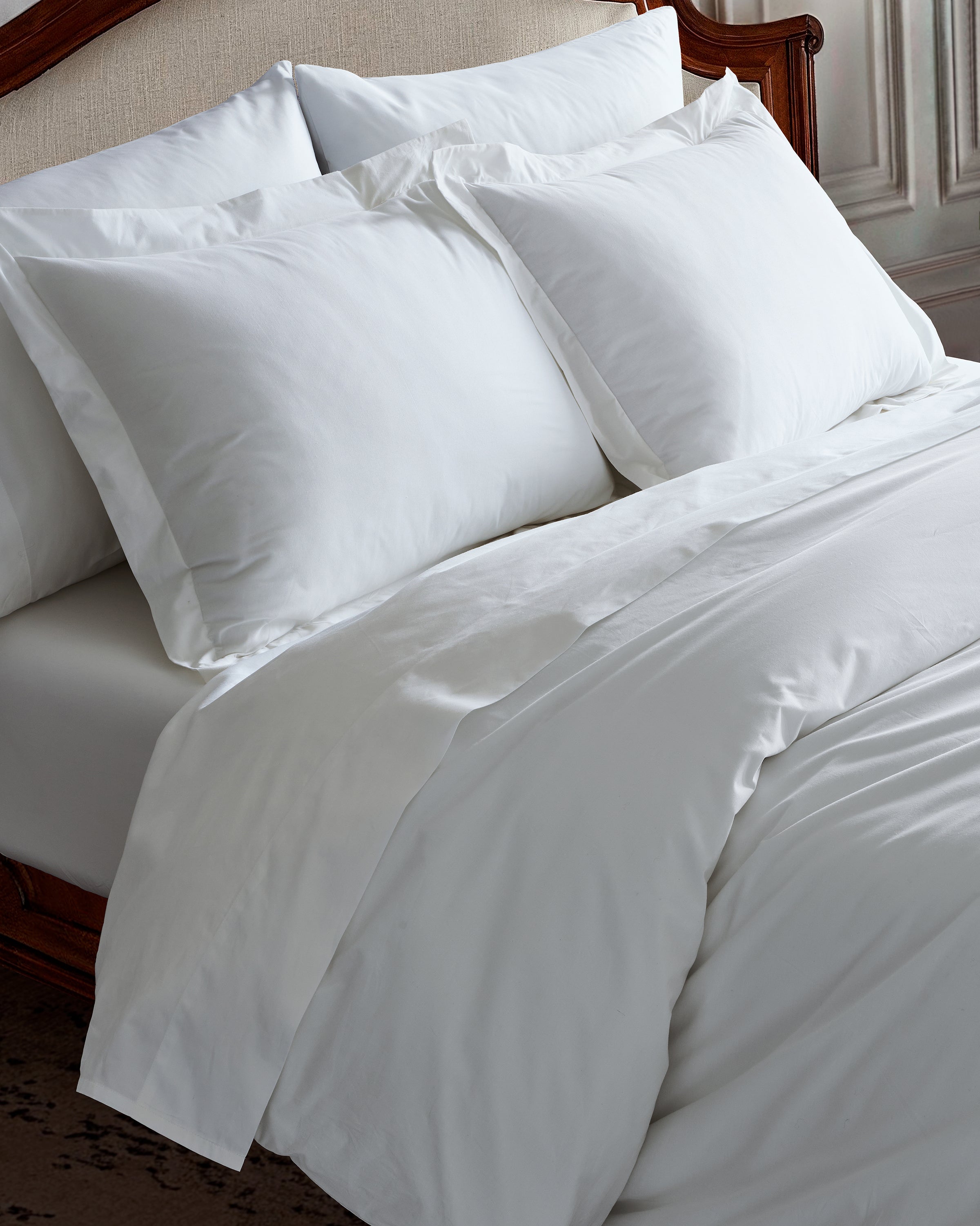 A neatly made bed with crisp 100% cotton sheets and fluffy white pillows against a wooden headboard exudes warmth and welcome, while the Luxe Premium Percale Duvet Set in White by Petite Plume enhances the comfortable and inviting appeal.