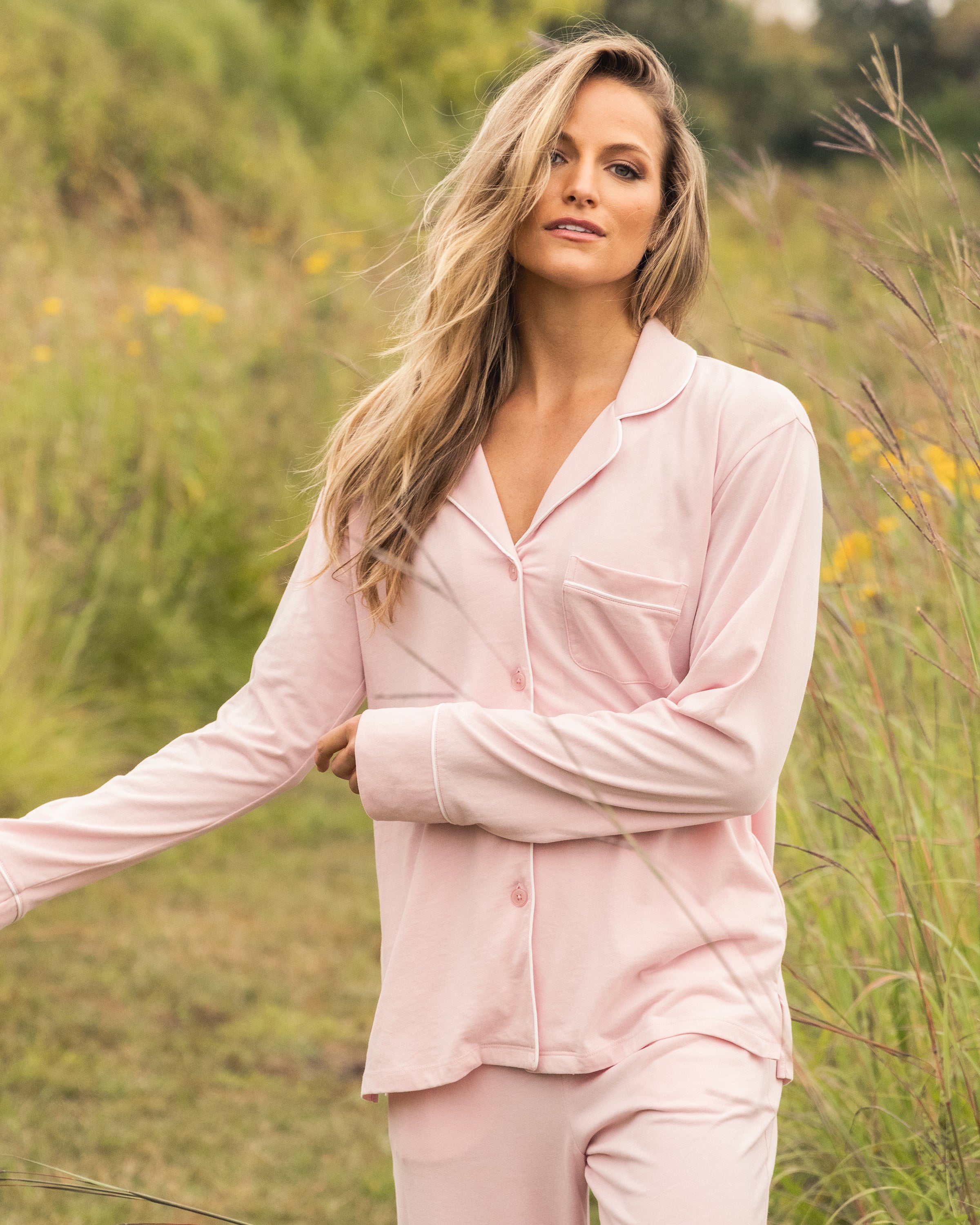 A person with long blonde hair stands outdoors in a grassy field, wearing the Womens Pima Pajama Set in Pink by Petite Plume. They smile at the camera amid lush greenery and yellow flowers, enhancing their luxury sleepwear vibe.