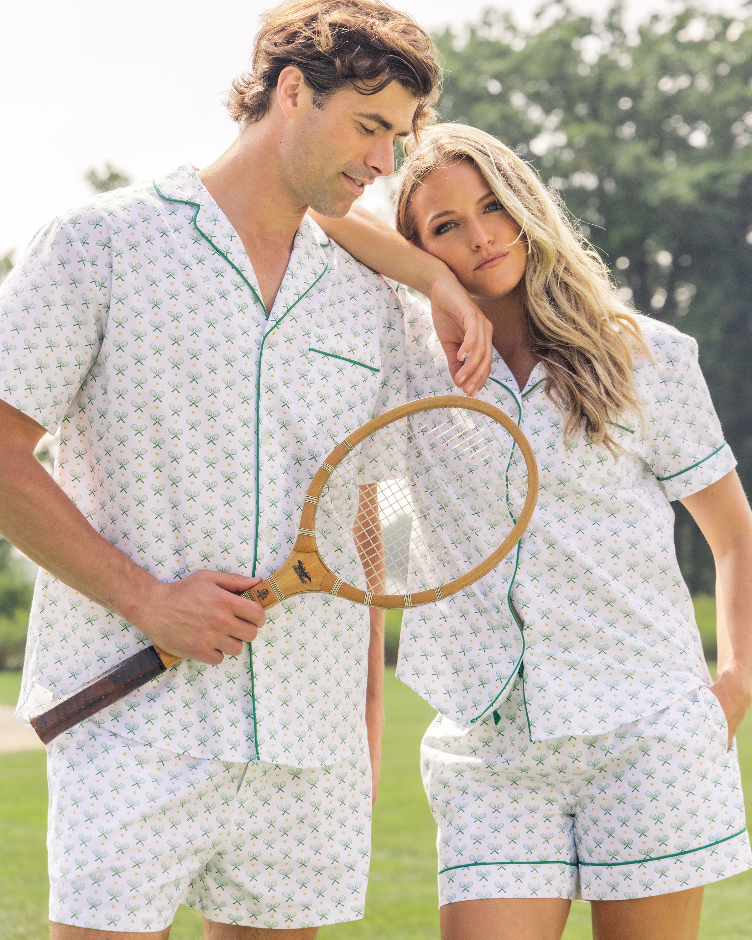 A man and woman stand outdoors in matching Petite Plume sleepwear, with the man wearing the Mens Twill Pajama Short Set in Match Point. The woman rests her head on his shoulder, holding a wooden tennis racket against a backdrop of lush greenery and towering trees.