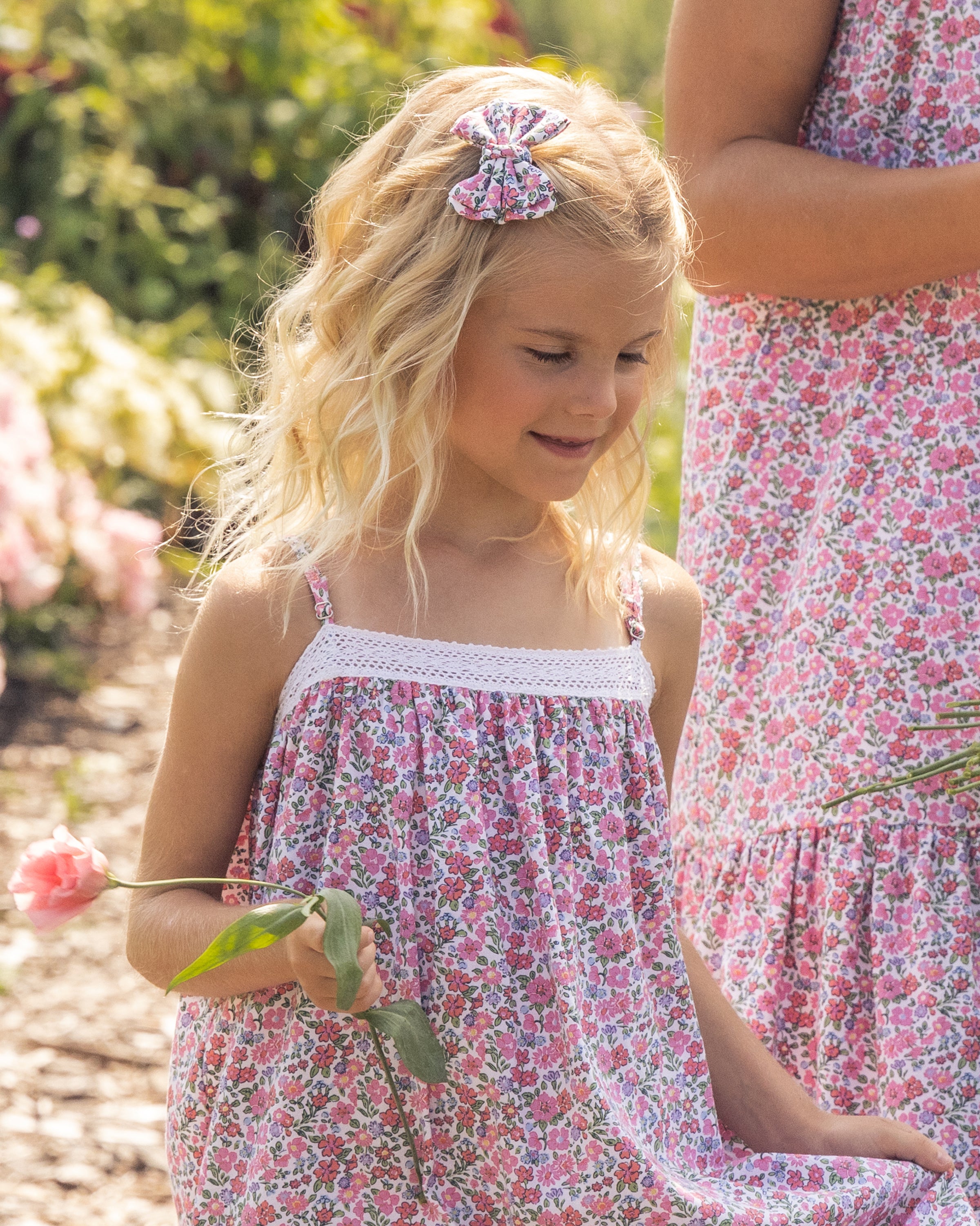 A curly-haired blonde girl wearing a Petite Plume pink floral dress with white lace trim and Fleurs de Rose hair bows holds a pink flower. Another person in a similar dress is partially seen beside her, surrounded by greenery and flowers outdoors.