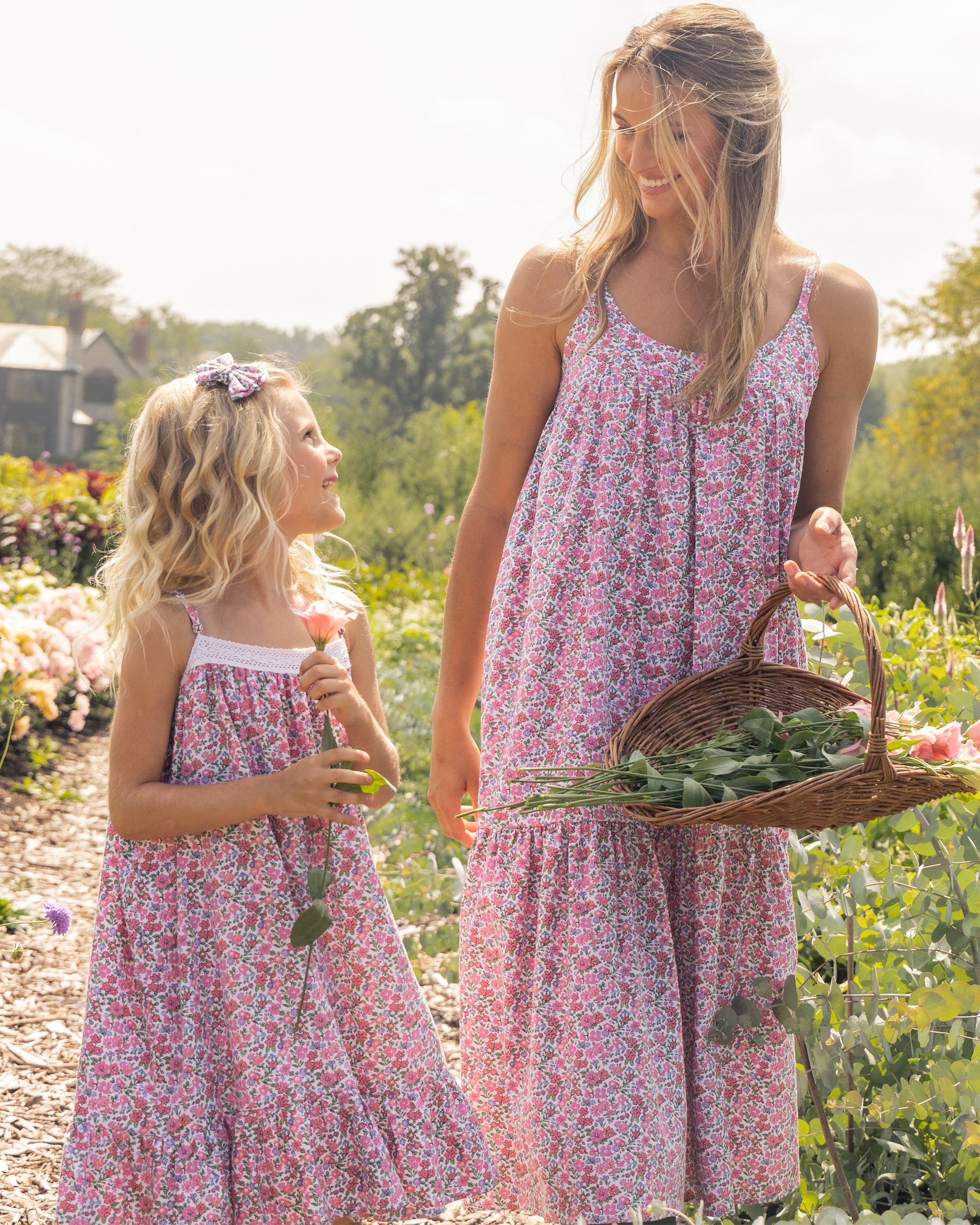 A woman in a Womens Twill Chloe Nightgown in Fleurs de Rose by Petite Plume strolls through a garden with a young girl, both basking in greenery. The woman holds a basket of leaves and flowers while the girl, clutching a blossom, smiles up at her against the scenic backdrop.