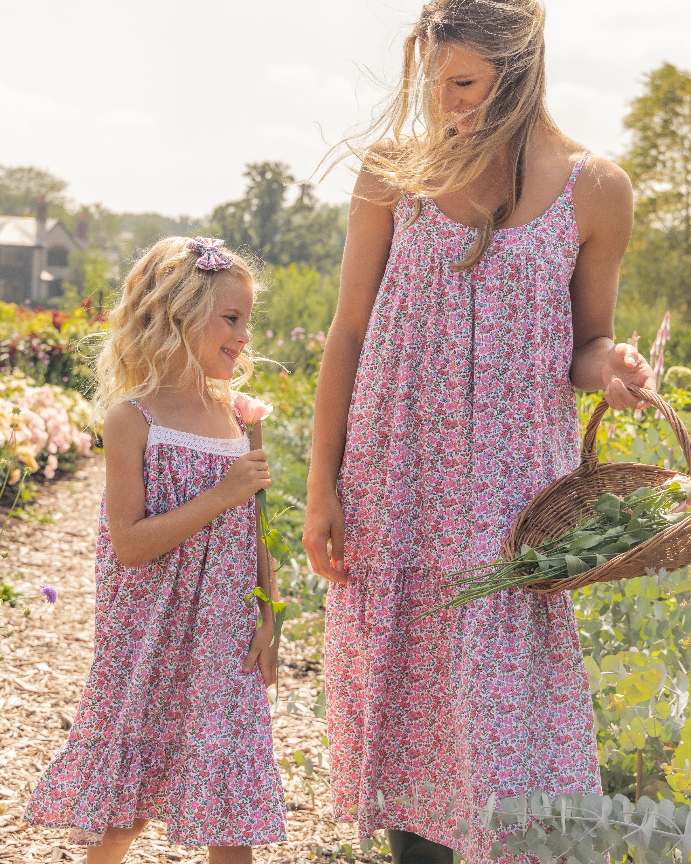 A woman and a girl in matching Petite Plume Fleurs de Rose print floral dresses walk down a garden path. The woman carries a basket of greens. They smile at each other, surrounded by blooming flowers under a partly cloudy sky.