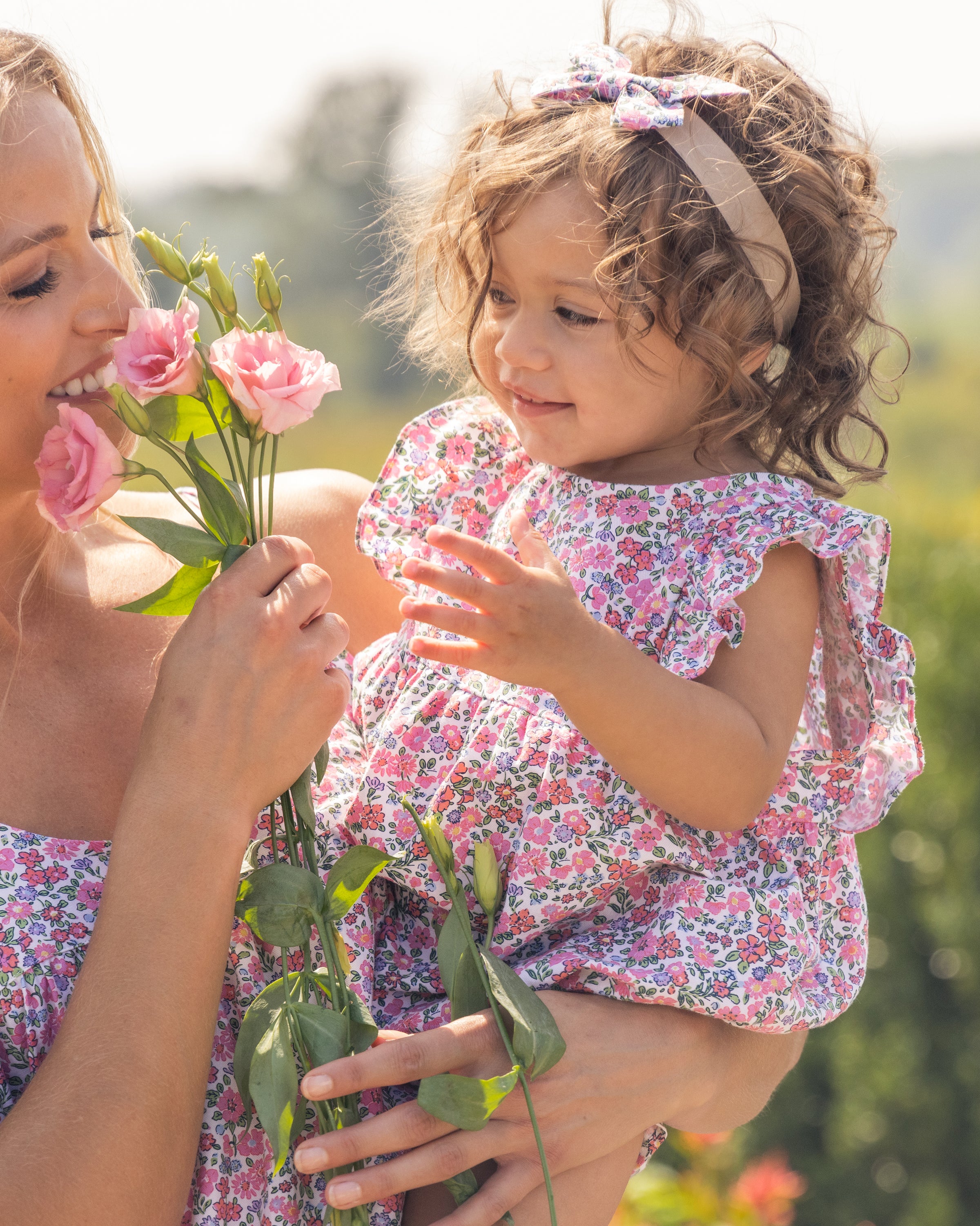 A woman and a curly-haired child in Petite Plumes Babys Twill Ruffled Romper in Fleurs de Rose enjoy the outdoors. The woman holds pink flowers, both admire, while the child wears a matching floral headband against a soft, green backdrop.