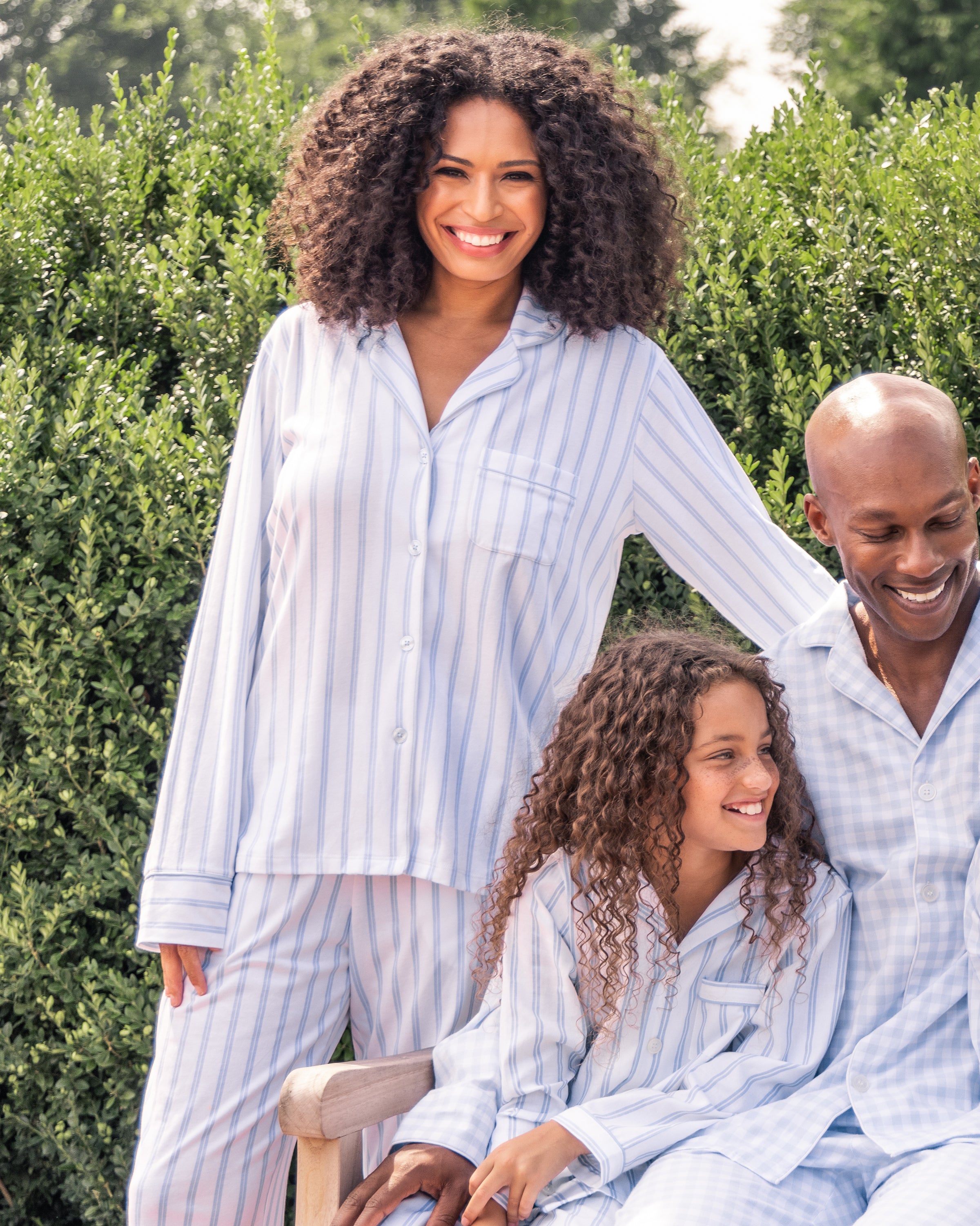A woman smiles outdoors in Petite Plumes Womens Pima Pajama Set in Periwinkle and White Stripe, matching her family as they relax together among green bushes. She rests her hand on the man’s shoulder while he and their child enjoy the luxurious sleepwear.