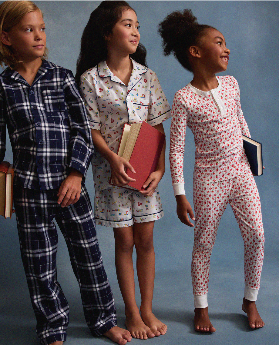Three children in pajamas stand barefoot against a blue background, each holding books. The child on the left wears dark plaid pajamas, the middle wears white printed short pajamas, and the right wears white pajamas with red patterns.