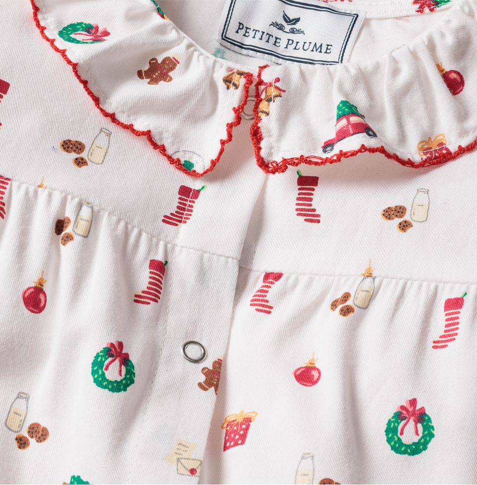 Close-up of a white childrens pajama top with red trim and festive holiday prints, including wreaths, stockings, gifts, gingerbread men, ornaments, milk, and cookies. The label reads Petite Plume.