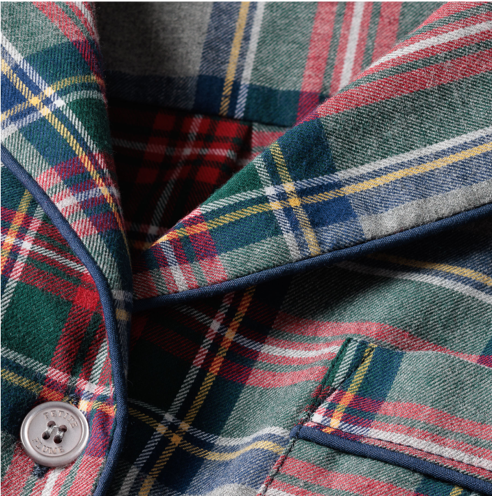 Close-up of a plaid fabric blazer or shirt in red, green, yellow, and navy blue with white and blue piped edges, a button, and part of a pocket visible.