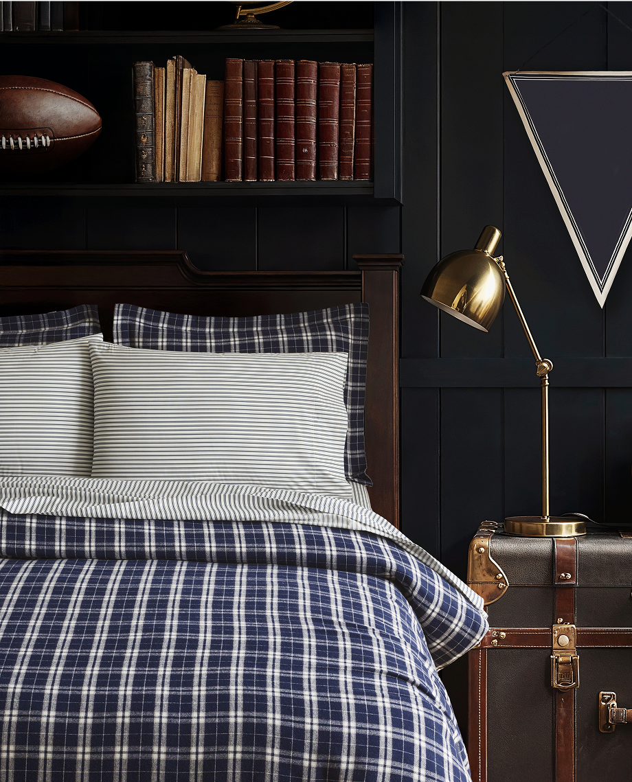 A neatly made bed with blue and white plaid bedding and striped pillows is in front of dark bookshelves, a gold desk lamp, and a vintage trunk. Books, a football, and triangular wall art are seen in the background.