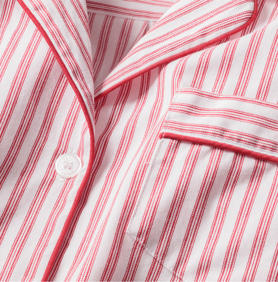 Close-up of pink and white striped fabric with a button, collar, and chest pocket, suggesting a classic pajama top. The stripes are evenly spaced, and the pocket has a matching striped pattern and piping detail.