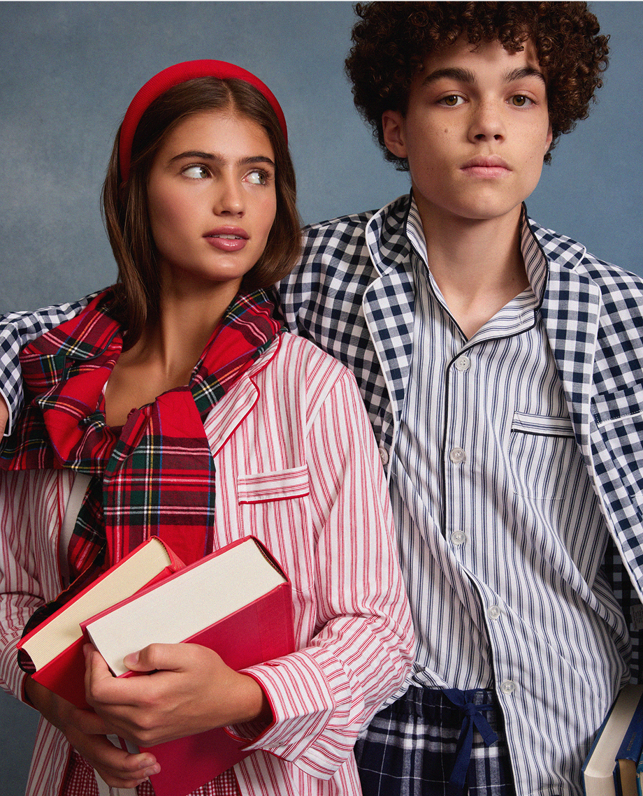 Two young people wearing striped and plaid pajamas stand side by side holding books. The young woman has a red headband and scarf, while the young man has curly hair and wears a plaid jacket over his pajamas.