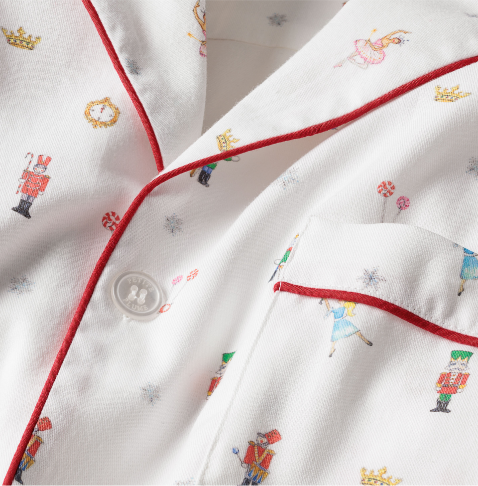 White pajama top with red piping and small printed Nutcracker-themed characters and objects, such as nutcrackers, ballerinas, candy canes, crowns, and snowflakes. A close-up view shows a pocket and button.
