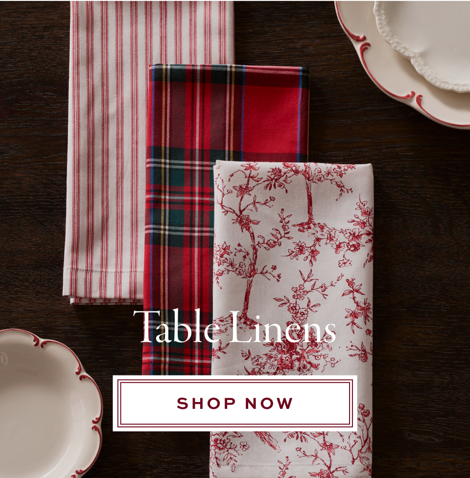 Three folded table linens—one with red stripes, one with a red and green plaid, and one with a red floral pattern—are displayed on a dark table with white plates. Text reads Table Linens, Shop Now.