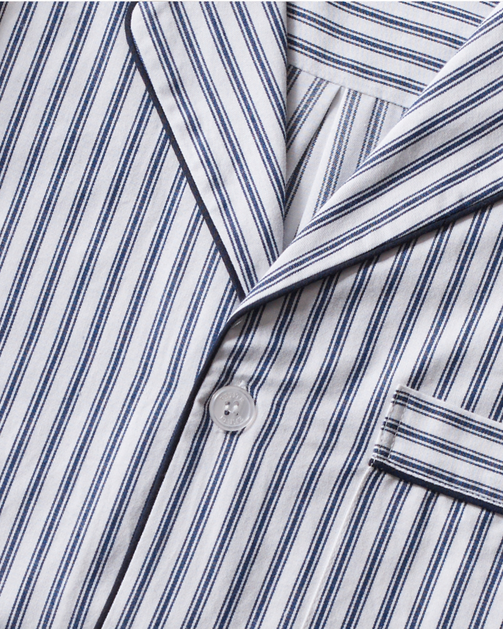Close-up of a classic pajama top with blue and white vertical stripes, featuring a notched collar, a white button, and a breast pocket with matching striped pattern and dark piping on the edges.