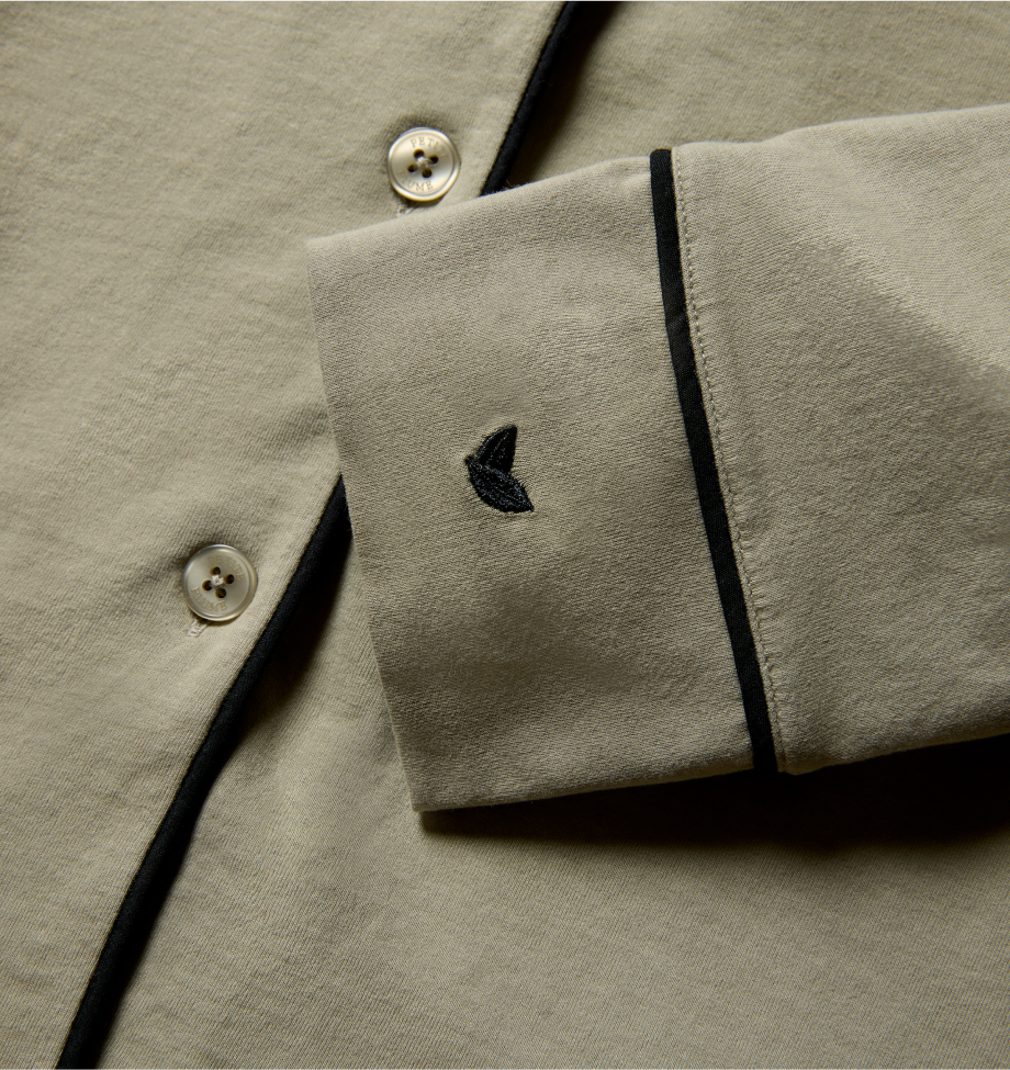 Close-up of a beige shirt with black piping along the edge, cream-colored buttons, and a small black embroidered logo on the cuff.