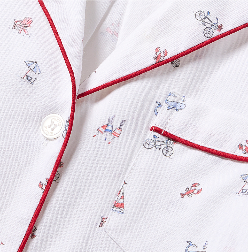 Close-up of a white shirt with small, colorful prints of beach-themed items, including a lobster, bicycle, sailboat, beach chair, and umbrella. The shirt has red piping and a white button near the pocket.