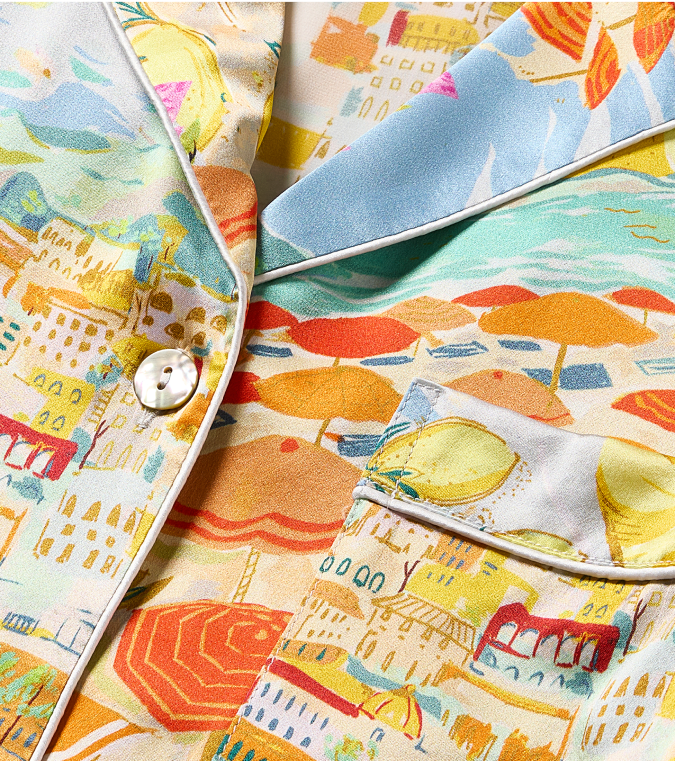 Close-up of a colorful, patterned fabric shirt with a pearl button and pocket, featuring illustrations of umbrellas, buildings, and seaside scenery in pastel tones.