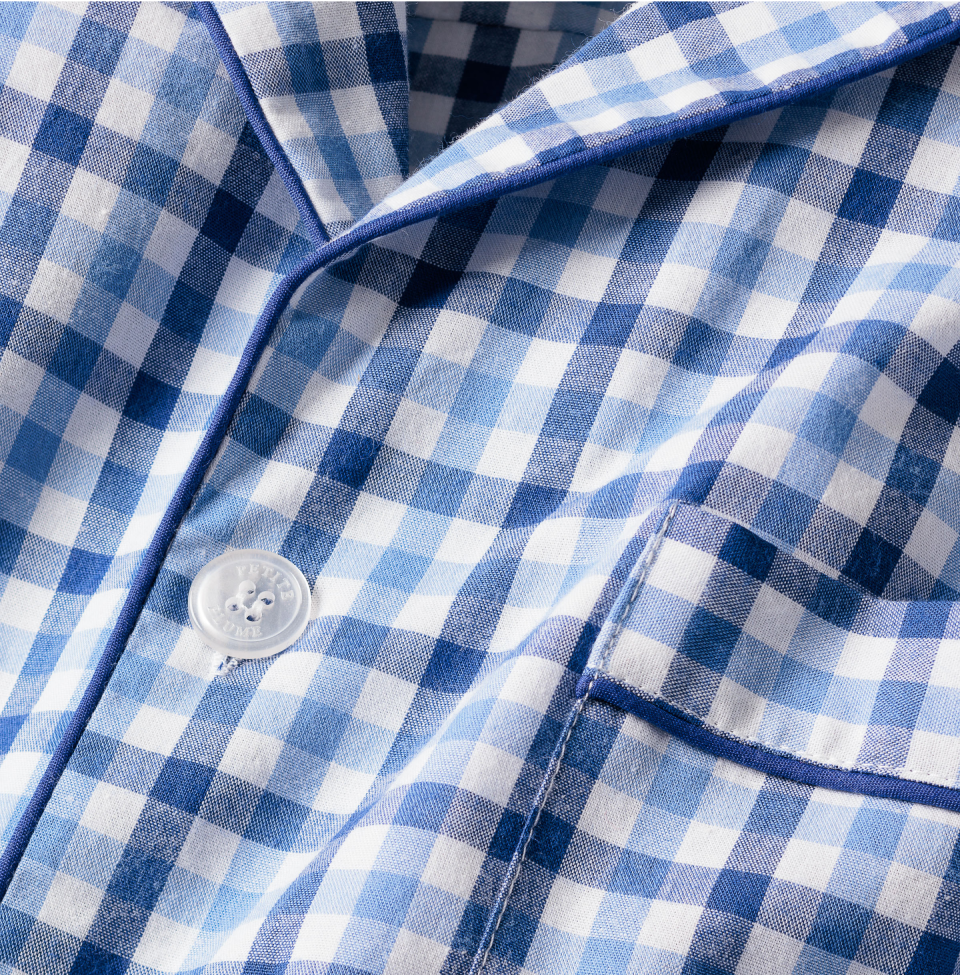 Close-up of a blue and white checkered fabric shirt with a button and part of a pocket visible, featuring dark blue piping along the edges.