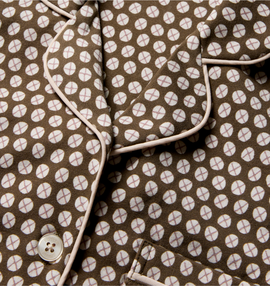 Close-up of a brown fabric with a repeating pattern of white circles crossed with red lines, featuring piping along the collar and a beige button near a pocket.