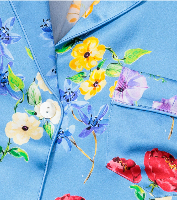 Close-up of a blue fabric shirt with a chest pocket, white buttons, and a colorful floral print featuring yellow, purple, red, and cream flowers with green leaves.