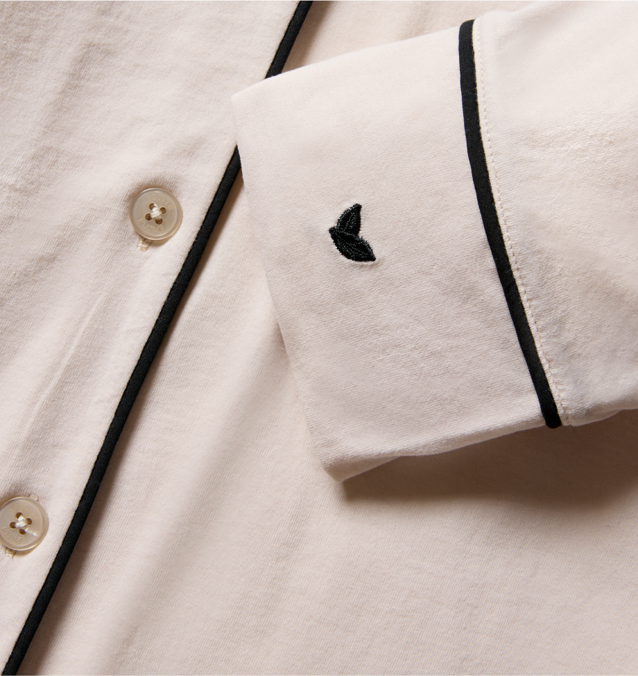 Close-up of a cream-colored shirt with black piping along the edge, white buttons, and a small black embroidered leaf on the cuff.