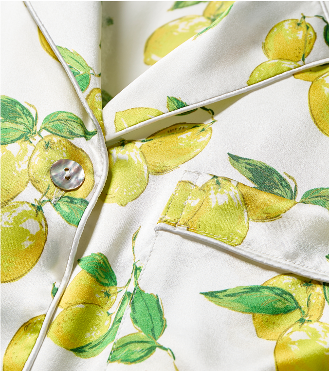 Close-up of a white fabric blazer featuring a vibrant yellow lemon and green leaf print, with a front pocket and a single shiny button visible.