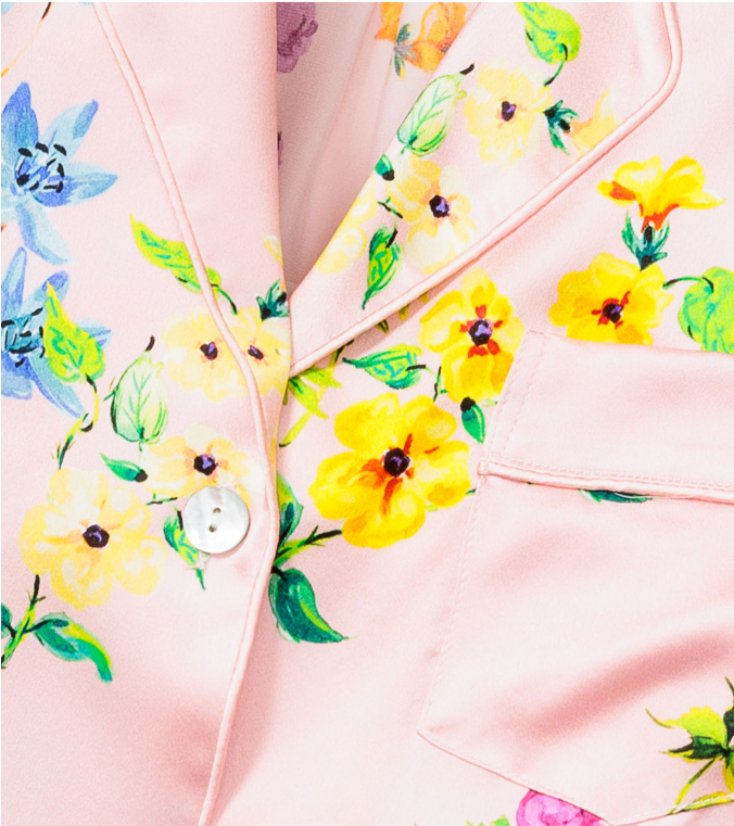 Close-up of a light pink blazer with vibrant yellow and purple floral prints, a white button, and a front pocket with pink piping detail.