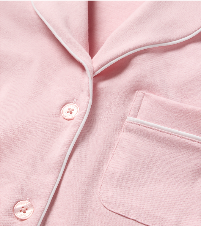 Close-up of a light pink pajama top featuring white piping along the collar, pocket, and button placket, with two pink buttons and a chest pocket visible.
