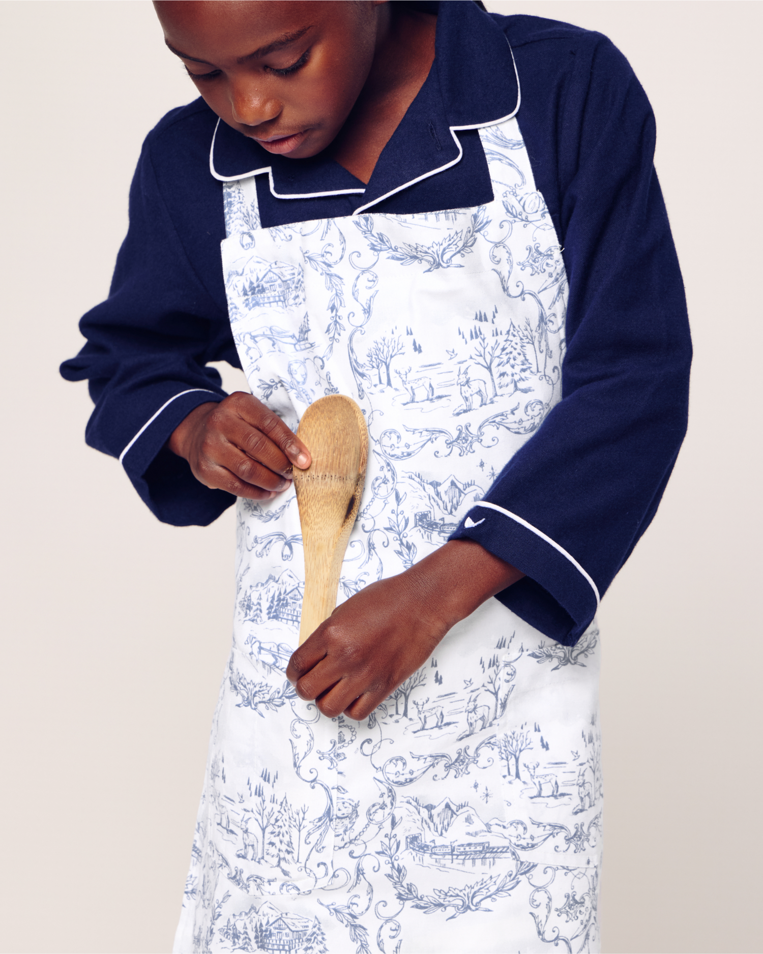 A child wearing navy blue pajamas and a white apron with blue patterns holds a wooden spoon, looking down as they prepare to cook or bake.