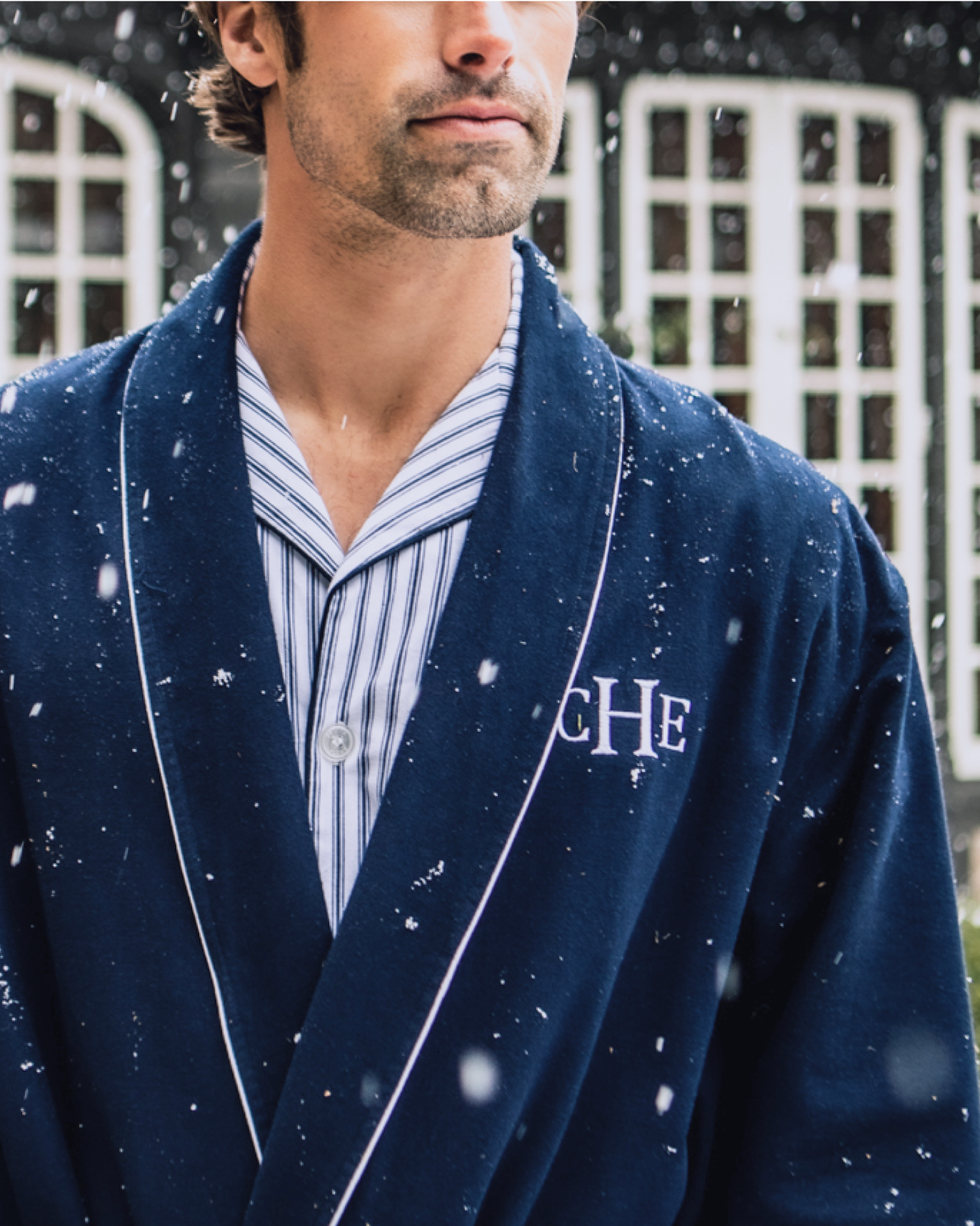 A man wearing a blue robe with white piping and a striped pajama top stands outside in falling snow. The robe has CHE embroidered on the chest, and the background shows arched windows.