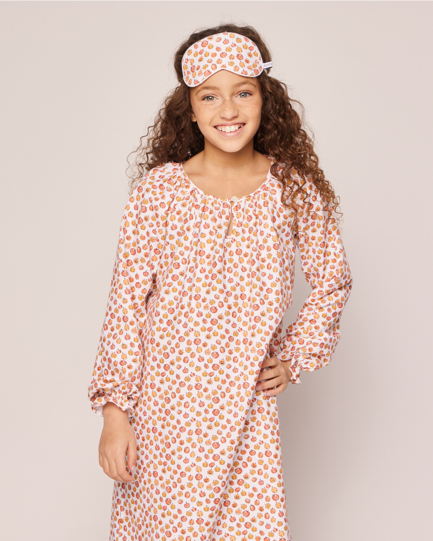 A smiling girl with curly hair wears a matching pink and orange patterned nightgown and sleep mask, posing with one hand on her hip against a plain light background.